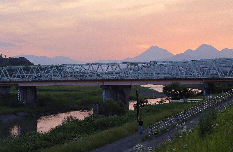 明磧橋が夕暮れの美しい風景の中に描かれています。明磧橋は鉄骨トラス構造の橋で、川をまたいで伸びています。両岸には緑豊かな木々が広がり、自然の美しさが際立っています。背景には山々がシルエットとなり、オレンジやピンクに染まる空と調和しています。橋へ続く道には歩いている人の姿も見え、スケール感と人の存在が加わっています。この光景は静かで、建築美と自然が融合した魅力的なものとなっています。