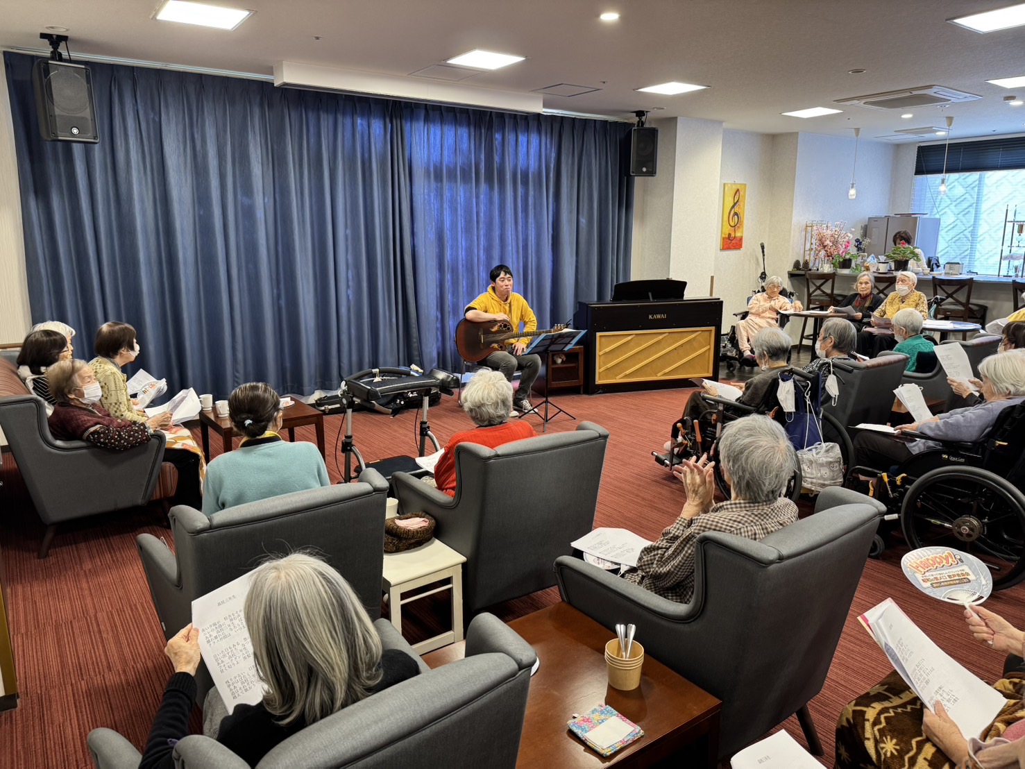 グランドホーム八雲で開催された音楽会の様子。プロのミュージシャンがギターを弾きながら歌い、高齢の観客が椅子に座って歌詞を見ながら楽しんでいる。室内にはピアノもあり、音楽会の温かい雰囲気が漂っている。