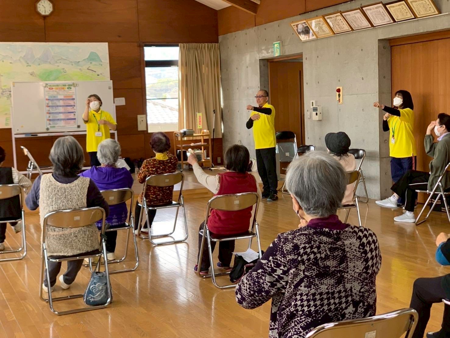 明磧町いきいきサロンの様子。参加者は椅子に座り、黄色いシャツを着たインストラクターの指示に従って腕を動かしている。インストラクターは前方に立ち、ホワイトボードには体操の手順が書かれている。壁には賞状や認定証が飾られ、室内には温かみのある雰囲気が広がる。参加者はマスクを着用しており、感染症対策が施されている