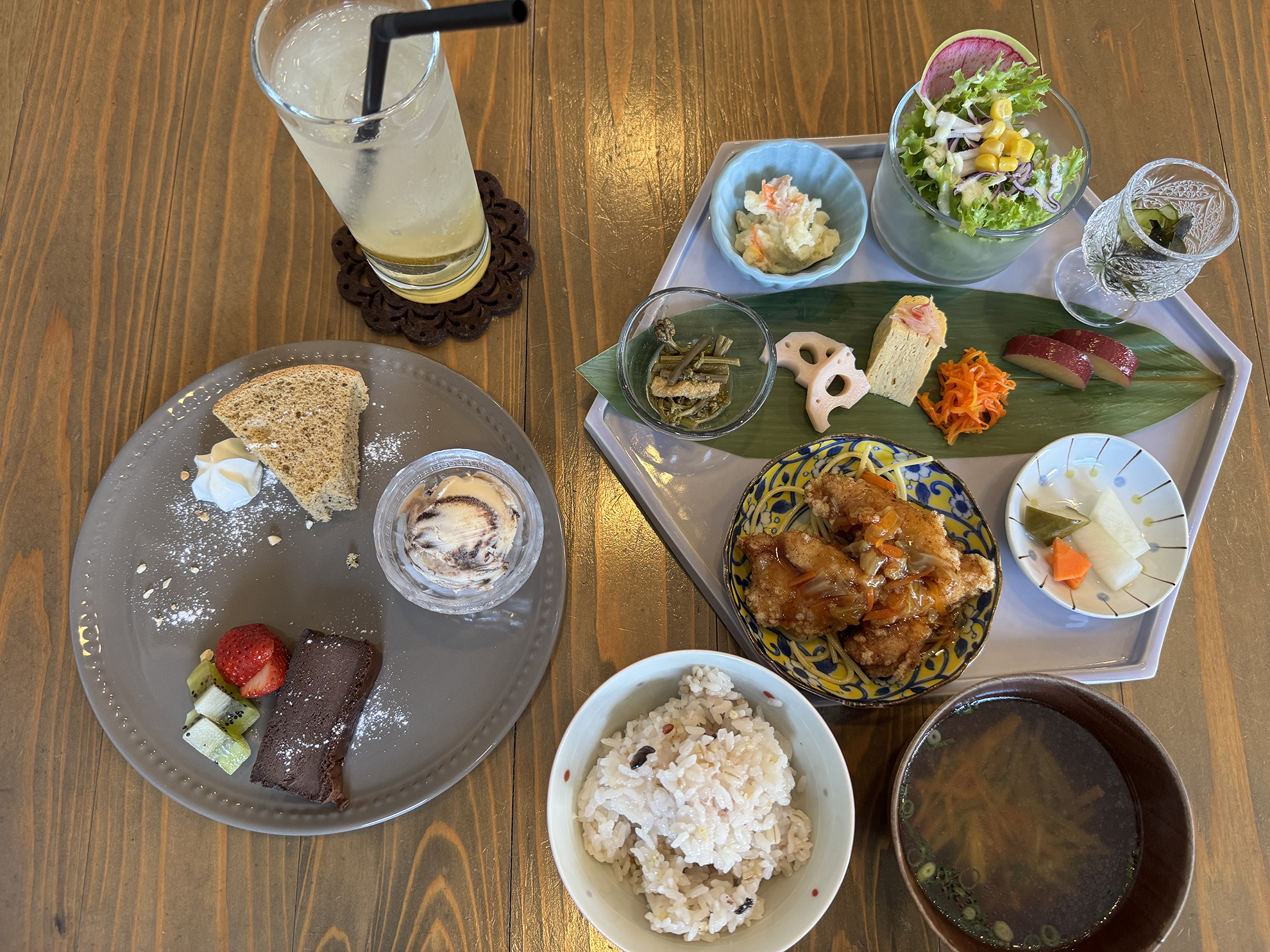 セットメニューが並べられています。左上に飲み物、右上のトレイにサラダ、ポテトサラダ、煮物、卵焼き、漬物、揚げ物など、右下に味噌汁、中央下に白米、左下のプレートにはデザートのケーキ、アイスクリーム、フルーツが盛り付けられています。