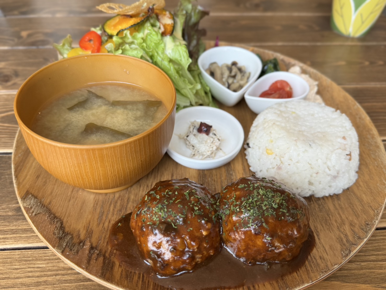 ハンバーグプレートランチ。木製プレートに盛り付けられた和風ランチセット。白ご飯、サラダ、味噌汁、2つのハンバーグ、小鉢料理3品が並び、彩り豊かでバランスの取れた食事が魅力。