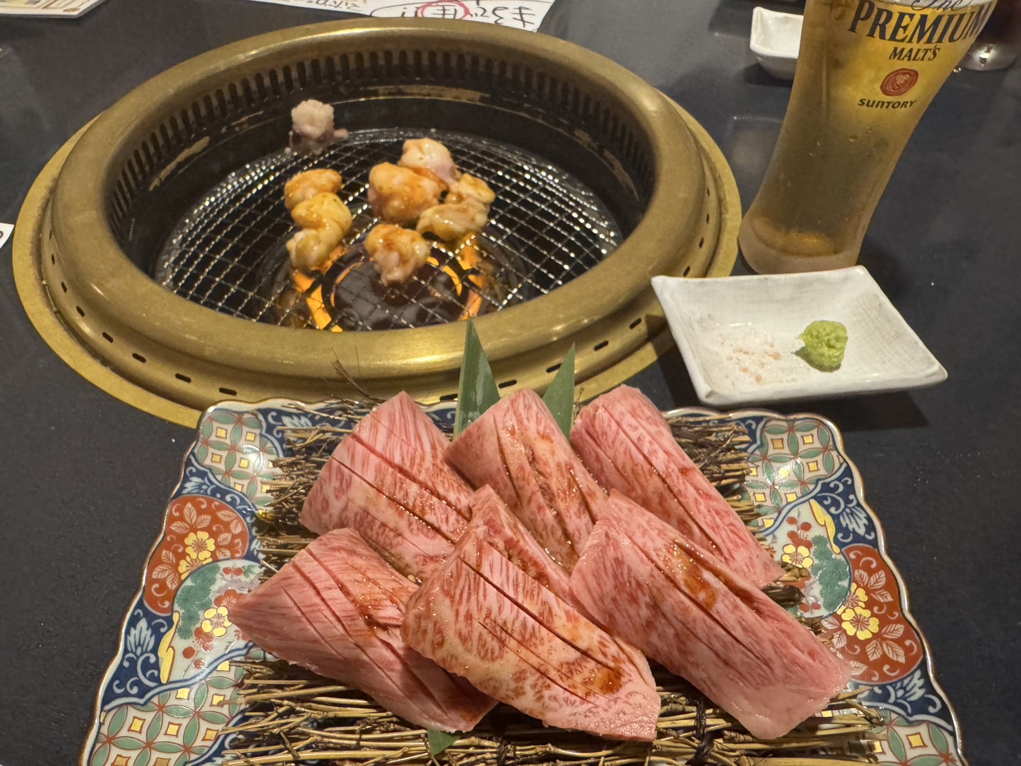 大分市上田町の焼肉MEAT KUNで提供される希少部位の霜降り肉が彩り豊かな皿に盛られ、炭火の焼き網でホルモンを焼いている様子。横にはビールとわさびが添えられている。