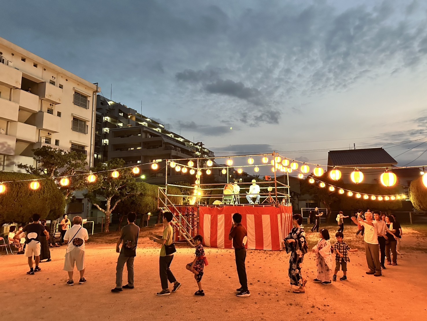 夕暮れ時の上田町で開催される盆踊り大会。赤白のやぐらを囲み、浴衣姿の人々が輪になって踊る。提灯が灯り、夏祭りの風情が漂う。