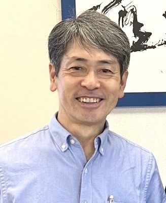 書道家・松本重幸さん
(城南在住)の顔写真。書をバックにさわやかな笑顔でこちらを見ています。