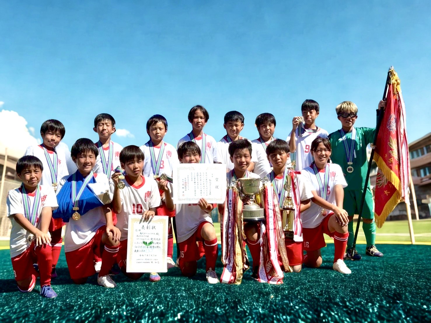 優勝した南大分サッカー少年団のみなさんの集合写真。１５人のチームメンバーが、賞状、優勝旗、優勝カップを掲げユニフォームを着ています。首から優勝メダルをそれぞれかけ、グランドで誇らしげな表情をしています。