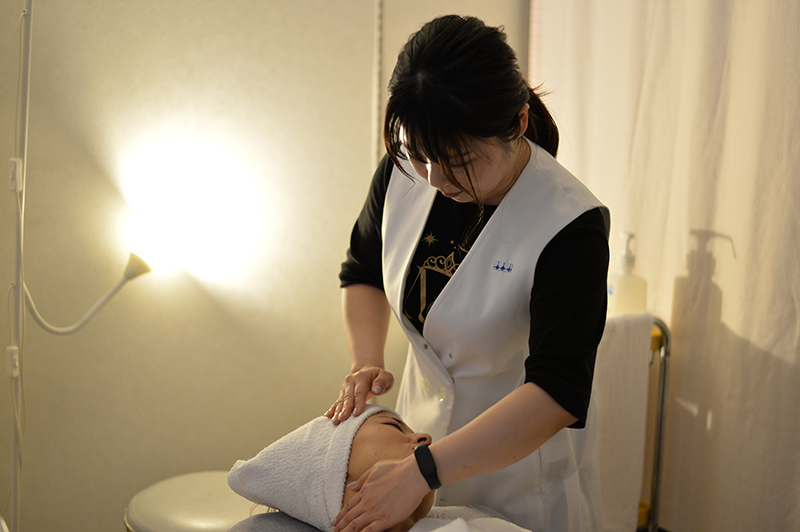 白衣を着た施術者(三浦 千尋さん)が、タオルを巻いたクライアントの顔を優しくマッサージしている一場面。暖かい照明と清潔感のある空間が、リラックスした雰囲気を演出している。