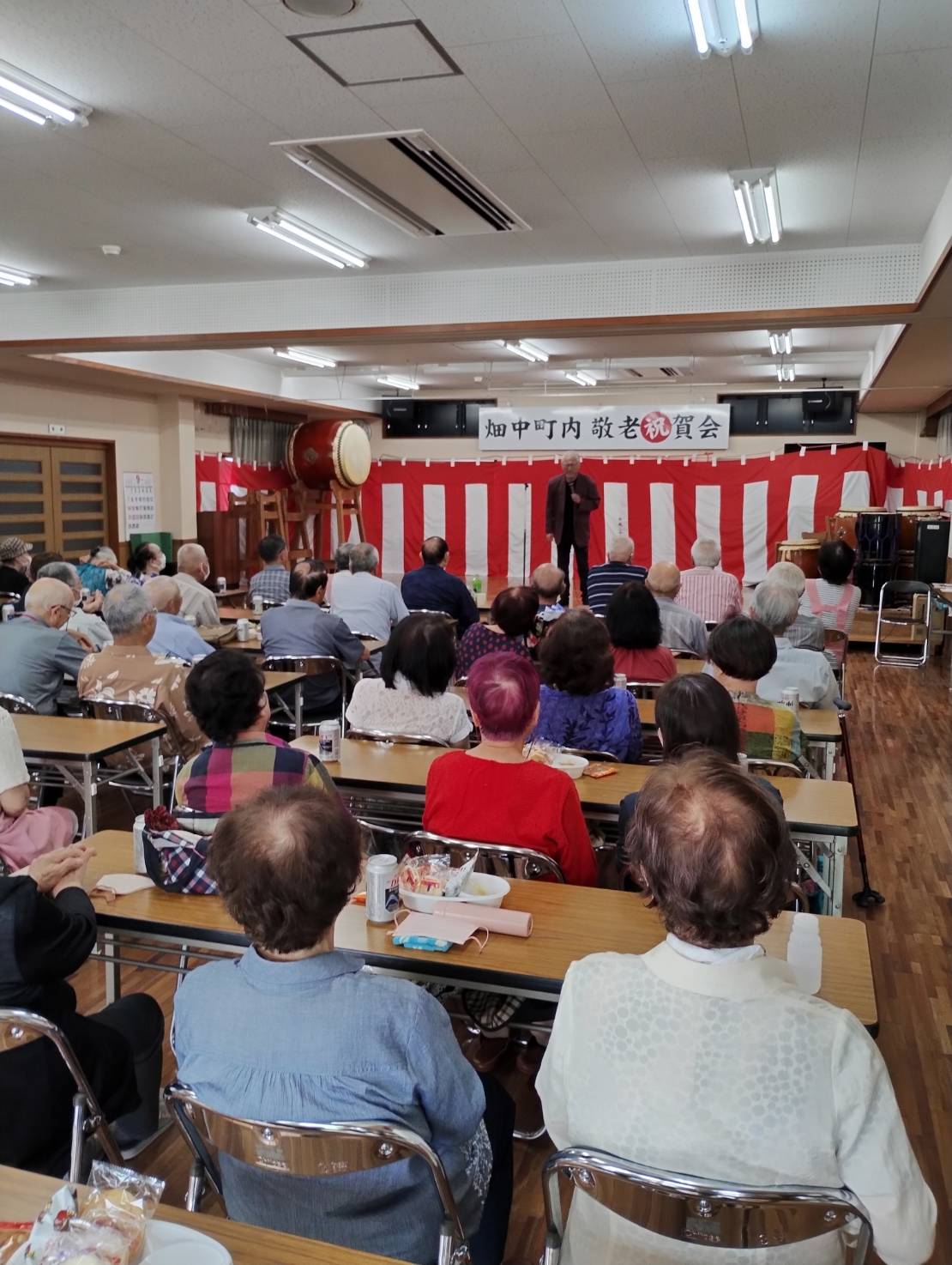 畑中町内敬老祝賀会の様子。屋内ホールで開催されています。高齢者たちがテーブルに座って舞台を見つめており、舞台では赤白の幕を背景に人物が話しています。左側には和太鼓が並んでいます。