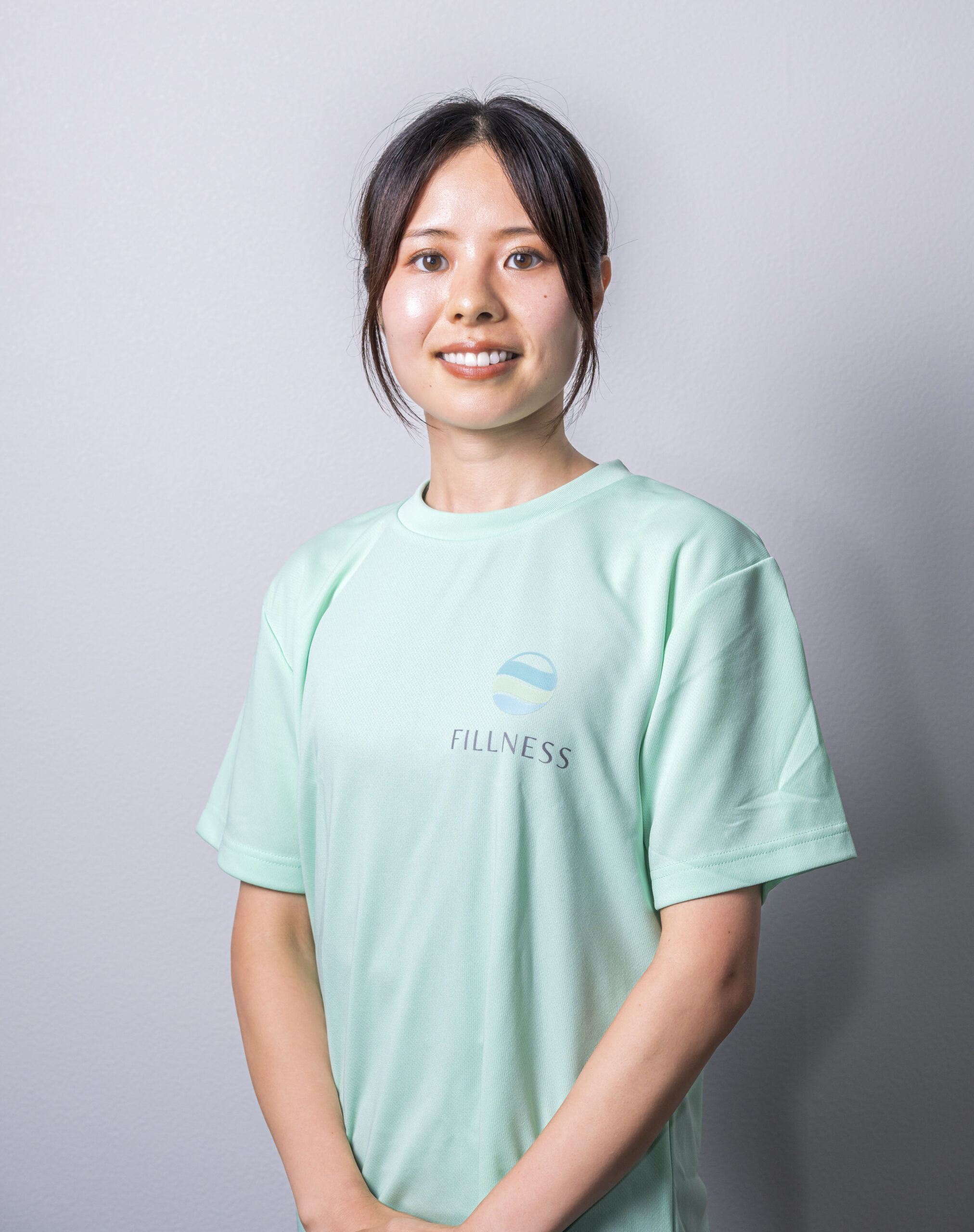 FILLNESS（フィルネス）ピラティスインストラクターの石田 花梨さん。ライトグリーンの布地にFILLNESSのロゴが入ったTシャツをきて笑顔でこちらを見ています。