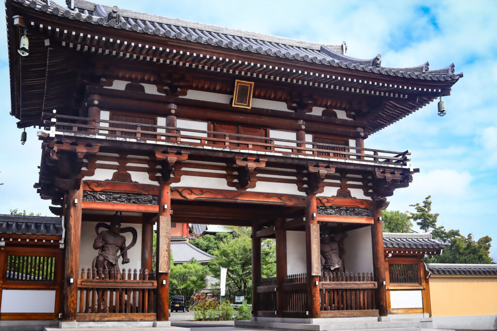 大分市の臨済寺にある総大門。木造の伝統的な寺院門で、反り上がった瓦屋根と精緻な梁の意匠が特徴。左右の龕には守護神像が安置され、荘厳な雰囲気を醸し出している。背景には境内の建物と緑が広がり、静謐な寺院空間を感じさせる。