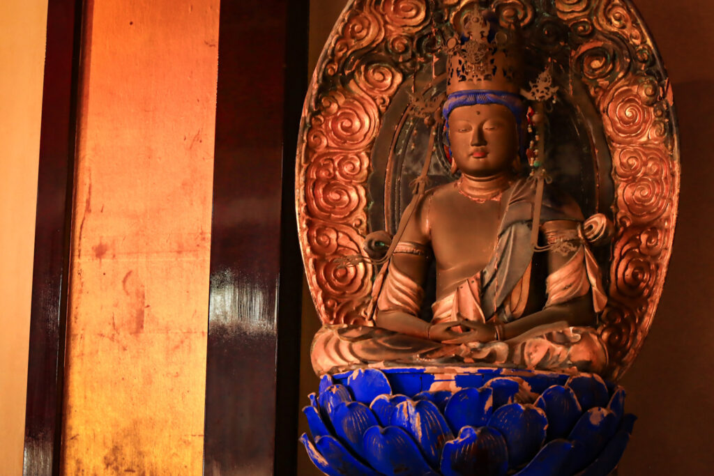 大分市・臨済寺に安置された釈迦如来坐像。華やかな冠と法衣をまとい、青い蓮華座に静かに坐す姿が印象的。両手は印を結び、背後には花文様の装飾が施された厨子が広がる。柔らかな照明が像の質感と彩色を際立たせ、敬虔な空気を漂わせている。