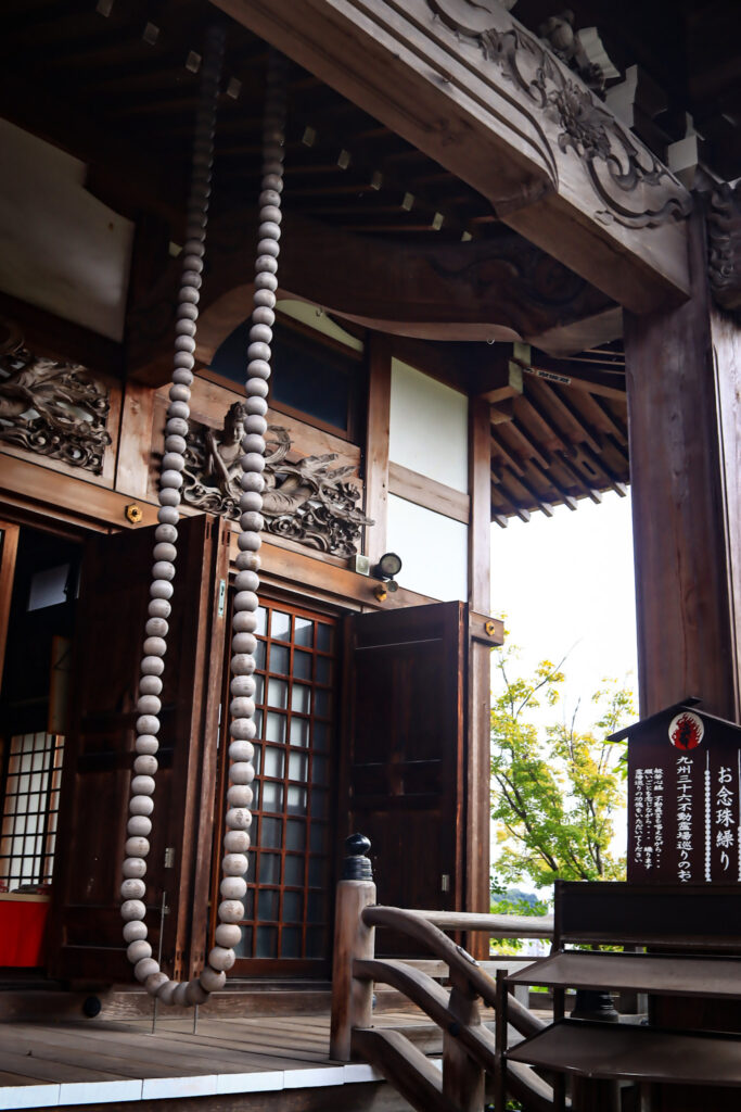 臨済寺本堂に吊るされたお念珠繰り。大粒の数珠が連なった縄が天井から垂れ下がり、参拝者が一粒ずつ撫でながら祈りを捧げるための仏具。木造の梁や格子戸、灯籠が周囲に調和し、静寂と敬虔さが漂う空間を演出している。