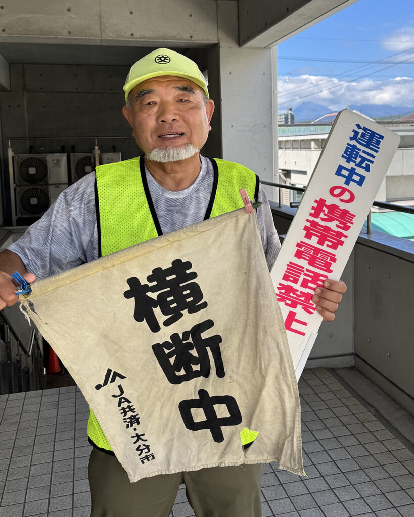 原 修一さんの写真。黄色い帽子をかぶり蛍光イエローのベストを着ています。手には横断中とJA共済・大分市と書かれた旗と運転中の携帯電話禁止と書かれた安全運転を呼び掛けるボードを持っています。