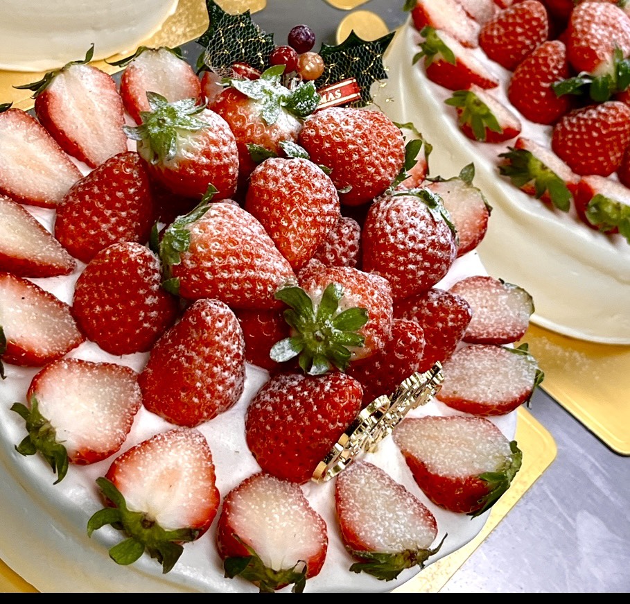 「ノエル・スペシャル」と名付けられたクリスマスケーキ。白いクリームの上に、丸ごとや半分にカットされた苺が円形に美しく並べられ、粉砂糖が雪のようにふりかけられている。中央にはヒイラギの飾りと「Merry Christmas」のプレートが添えられ、華やかで祝祭感あふれる仕上がり。金色の台座に載せられ、背景にも同様のケーキが並ぶ。