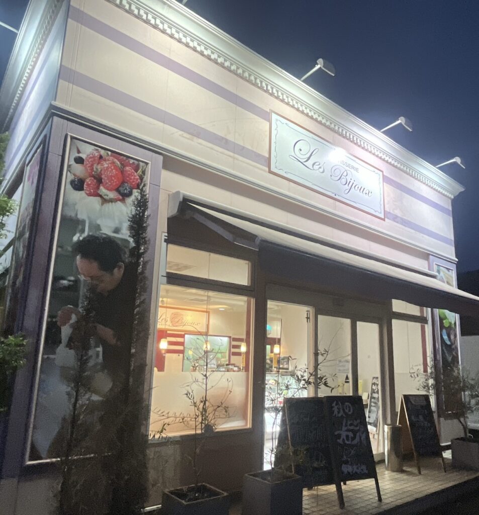 夜の街角に佇むケーキ店「パティスリーレビジュ」の外観。パステルカラーの横縞が彩るファサードに、店名のライトサインが温かく輝く。大きな窓からは明るい店内が見え、焼き菓子やスイーツが並ぶ様子がうかがえる。店先には観葉植物と手書きの黒板メニューが並び、左側のバナーには苺のデザートと調理中の人物の写真が添えられている。洗練された可愛らしさと親しみやすさが同居する、街の小さな宝石箱のような一軒。