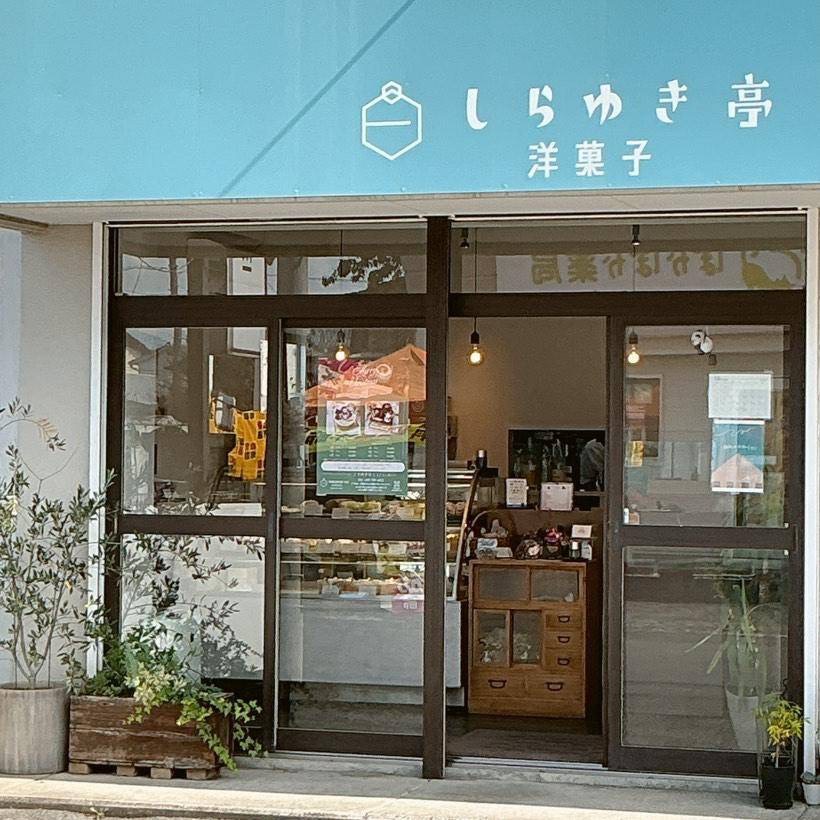 洋菓子店「しらゆき亭」の外観。青地に白文字の看板には、ケーキのロゴと「しらゆき亭」「洋菓子」の文字が描かれている。ガラス扉と窓からは、店内のケーキショーケースや温かみのある照明が見え、木製キャビネットが入口付近に配置されている。外には鉢植えが並び、親しみやすく清潔感のある雰囲気が漂う。