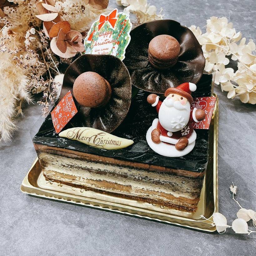 「ショコラノエル」と名付けられたクリスマス仕様のチョコレートケーキ。艶やかなチョコレートグレーズで覆われた層状のケーキの上に、花のような形のチョコレート飾りとマカロンが配置され、中央にはサンタの砂糖菓子と「Happy Christmas」「Merry Christmas」のプレートが添えられている。金色のトレーに載せられ、背景にはドライフラワーがあしらわれた、上品で祝祭感あふれる一枚。