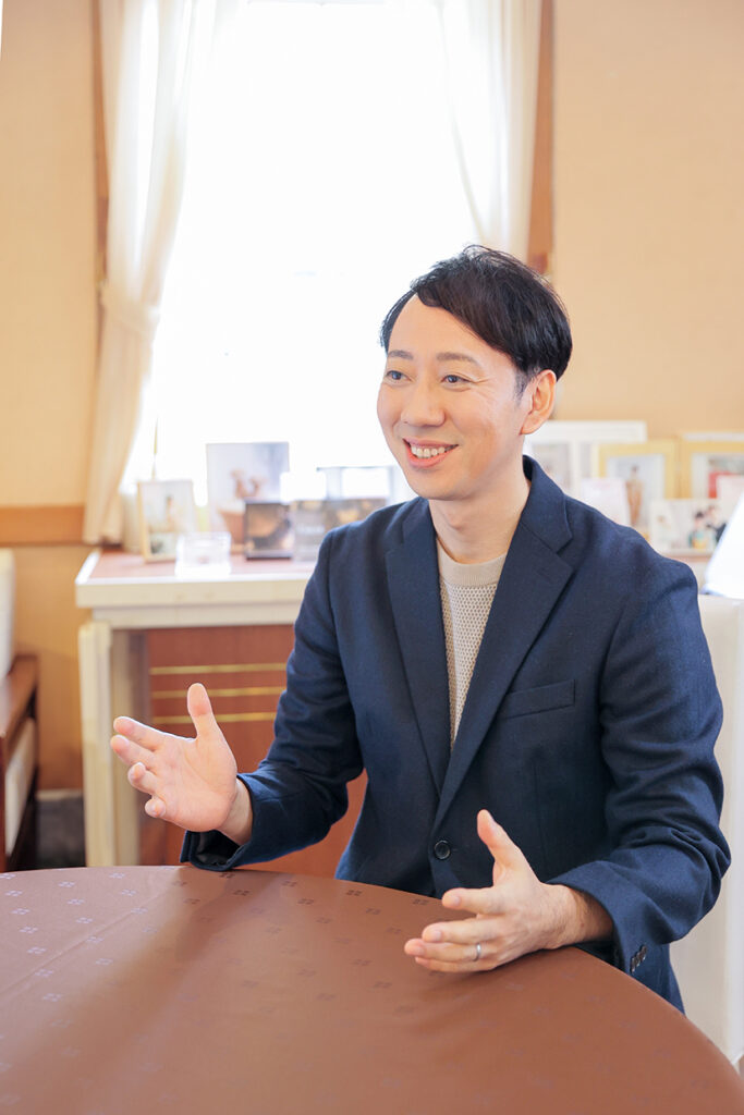 「結婚サポートルーム HOP STEP えんむすび」の田形博士さんが、丸テーブルを囲んで両手を使って語りかける様子。ダークブルーのジャケットとメッシュ調のインナーを着用し、穏やかな室内で対話を重ねる姿が印象的。背景には自然光が差し込む窓と、写真や書類が並ぶ棚が見え、親しみやすく信頼感のある雰囲気が伝わる。