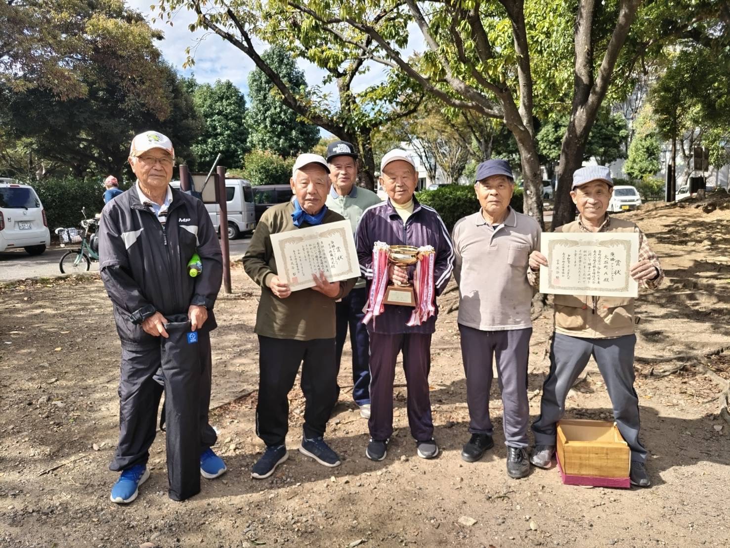 南大分体育協会主催「秋季球技大会・グラウンドゴルフ大会」の表彰風景。屋外の明るい空の下、優勝した大石町の皆さんが6人で並び、中央の人物は赤白のリボンが付いたトロフィーを掲げている。両脇の参加者は賞状を手にしており、達成感と仲間との喜びが伝わる一枚。背景には木々や車両が見え、地域のスポーツイベントらしい穏やかな雰囲気が漂っている。