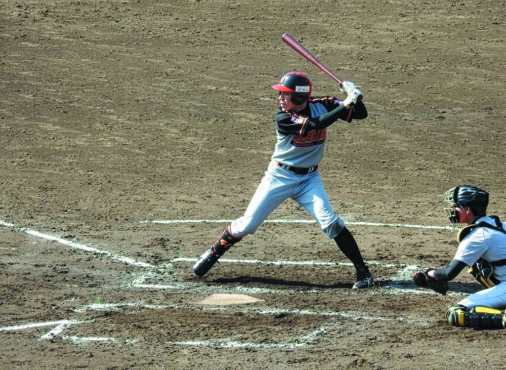 BAN BASEBALL CLUBの試合中の一場面。打席に立つ選手がピッチを待ち構え、グレーと黒を基調にオレンジのアクセントが入ったユニフォームとヘルメットを着用している。背後ではキャッチャーが防具を身に着けて構え、白いチョークラインが引かれた土のグラウンドが緊張感ある瞬間を際立たせている。打者と捕手の集中した姿勢が、競技の迫力とチームの真剣さを伝える一枚。