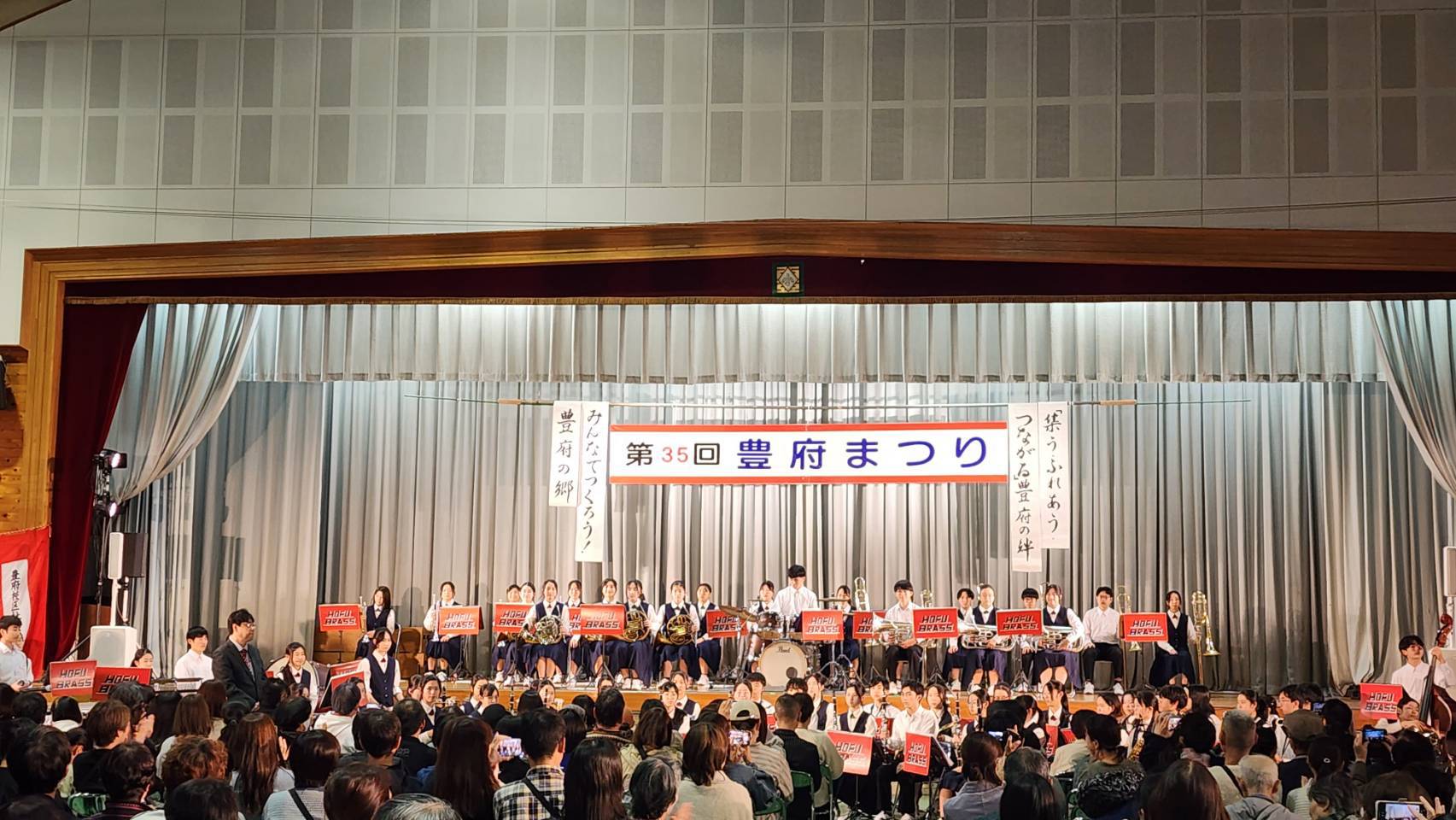 第35回豊府まつりのフィナーレで、大分豊府中高等学校吹奏楽部がステージ演奏を披露する様子。多数の部員が「HOFU BRASS」と書かれた赤いボードを掲げながら演奏し、観客の前で華やかに締めくくる。ステージ背面には「みんなでつくろう！豊府の郷」「集う・ふれあう・つながる豊府の絆」と書かれた垂れ幕が掲げられ、地域の絆と文化の誇りが感じられる場面。
