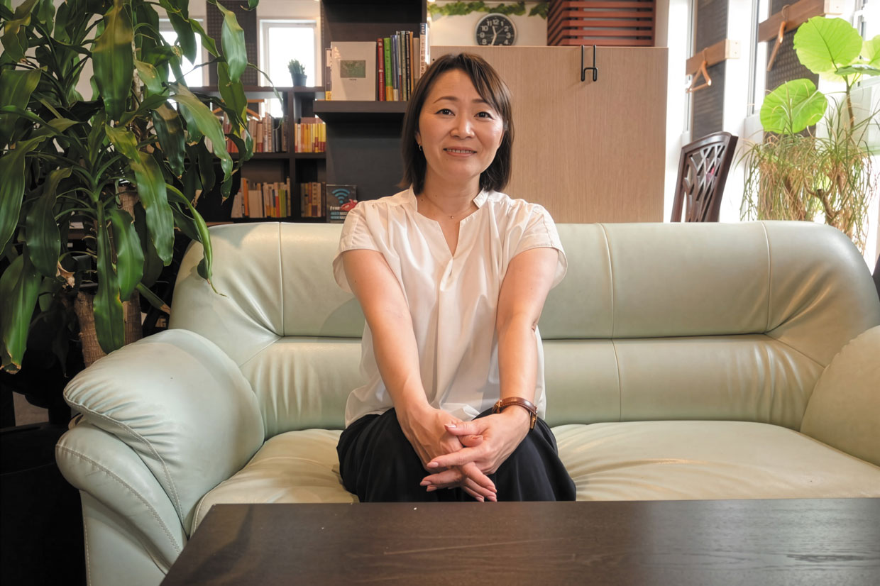 株式会社Bambitzの代表取締役・花田亜弥さんが、明るく落ち着いた雰囲気の室内でライトグリーンの革張りソファに座っている様子。白いブラウスとダークカラーのパンツを着用し、両手を膝の上で穏やかに組んでいる。背後には本棚や観葉植物があり、自然光が差し込む窓辺が空間に温かみを添えている。知性と親しみやすさが感じられる一枚。