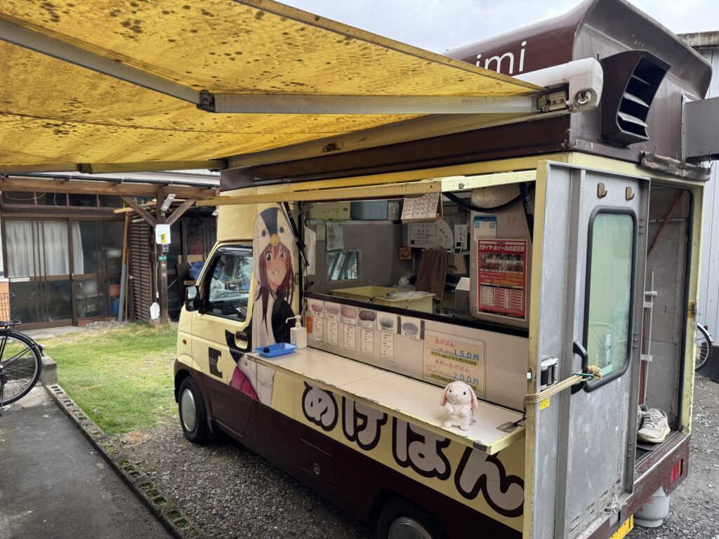 花園秋祭りの屋外風景。住宅地に停車した黄色と茶色のキッチンカーには、アニメ風キャラクターのイラストが描かれており、カウンターにはぬいぐるみと手書きのメニューが並ぶ。「あげぱん」や「あげぱんアイス」などの品目が掲示され、きなこ・ココア・カレーなどの味が選べる。車体の上には年季の入った黄色いオーニングが広がり、背景には和風の建物と自転車が見える。地域の温もりとポップカルチャーが融合した、親しみやすい屋台のひとコマ。