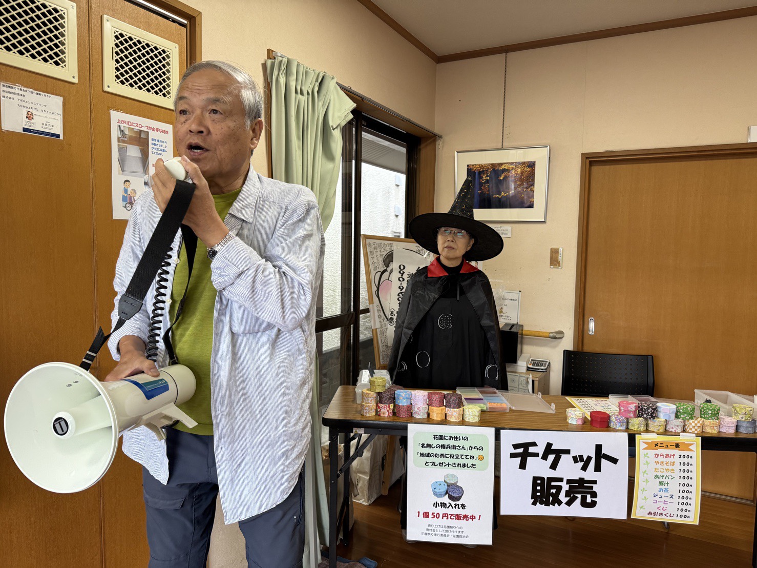 花園公民館で開催された「花園秋祭り」の一幕。左側では男性がメガホンで呼びかけを行い、右側には黒い帽子とマントをまとった魔女の仮装をした人物が、カラフルな容器が並ぶチケット販売ブースの後ろに立っている。テーブルには抽選券や小物販売の案内、焼きそば・たこ焼きなどのメニュー表が掲示され、地域の温かみと季節のにぎわいが感じられる。