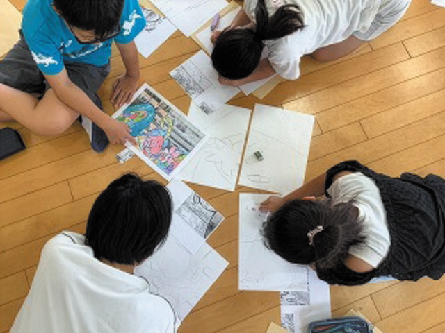 南大分小学校150周年記念式典の一環として行われた「ビックアートづくり」の様子。木の床に座った児童たちが、印刷された参考画像やスケッチ用紙を囲みながら、鉛筆で真剣に絵を描いている。互いに意見を交わしながら創作に取り組む姿が、協力と創造の楽しさを伝える。記念事業として、学校の節目を彩るアート制作の一場面。