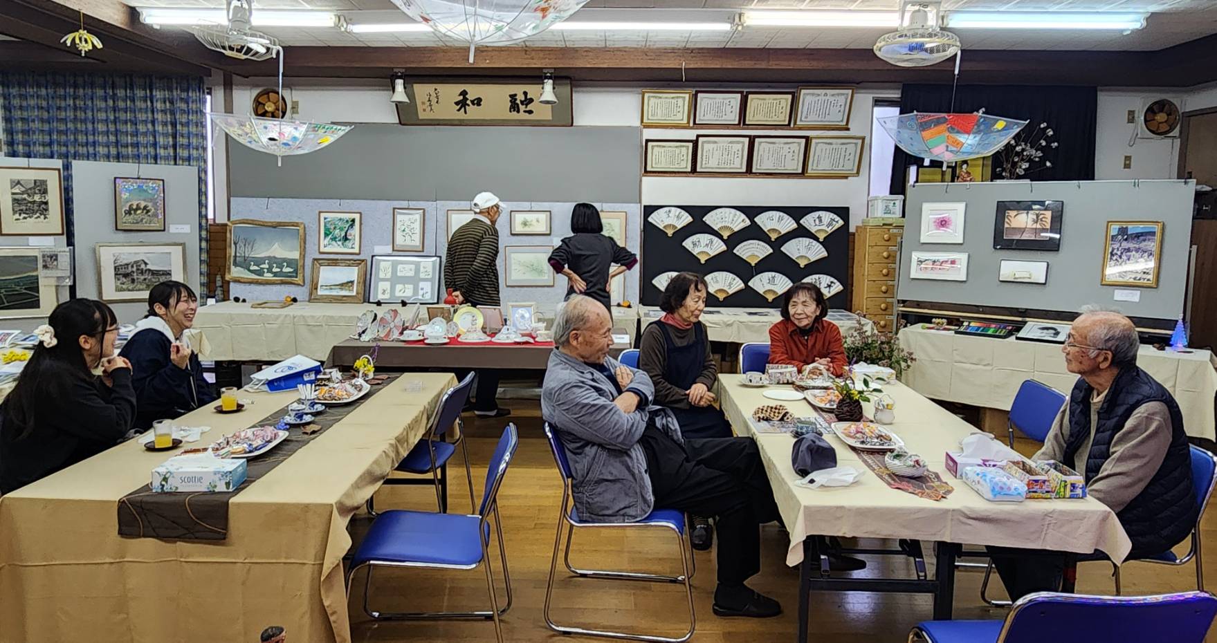 南太平寺公民館で開催された「我が家の自慢の一品展」の会場風景。絵画や書、飾り扇など多彩な作品が壁や展示パネルに並び、来場者が歓談しながら鑑賞している。テーブルには飲み物やお菓子が用意され、世代を超えた交流と作品への温かなまなざしが広がる、地域の文化とふれあいを感じるひととき。
