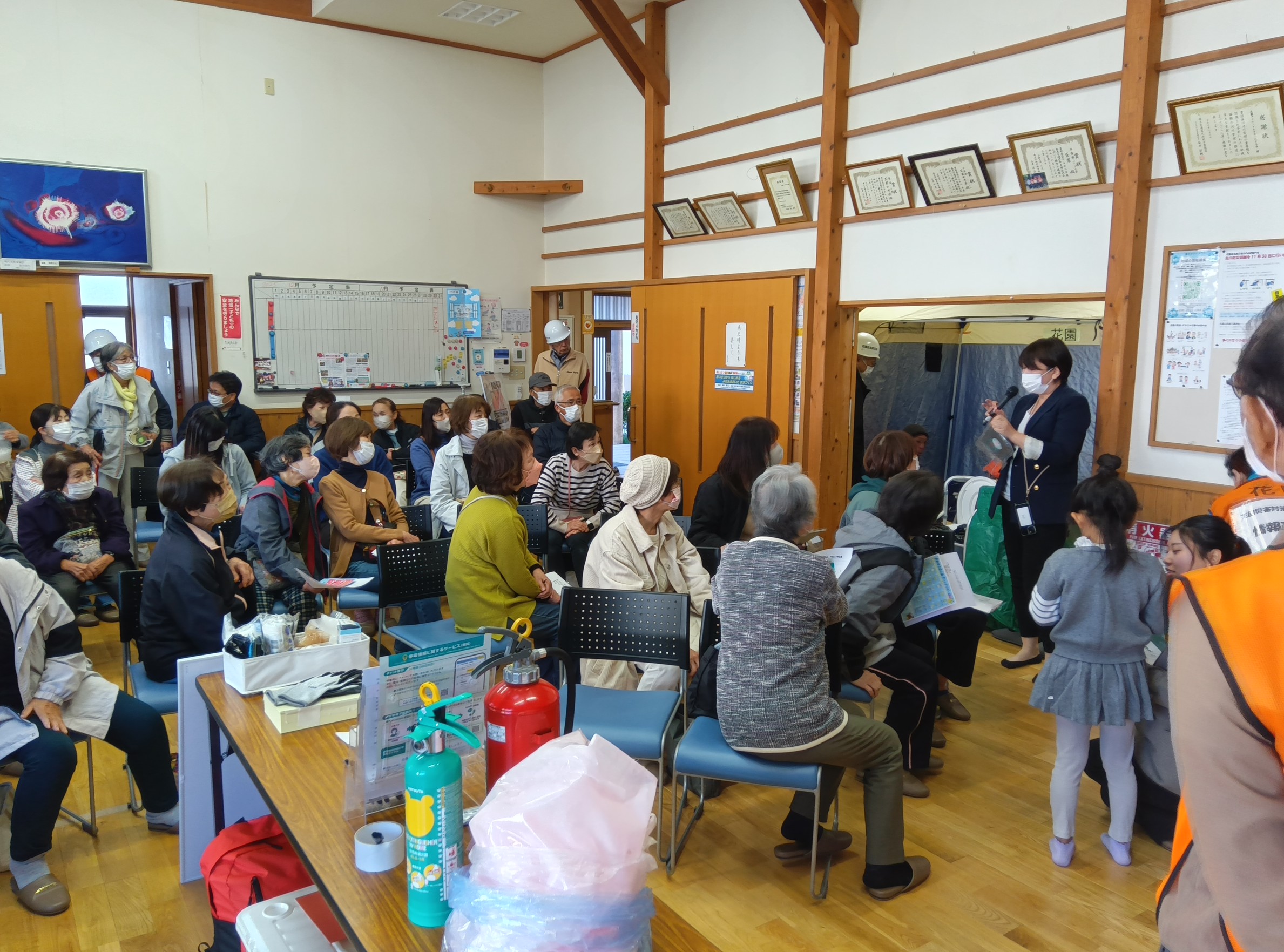 花園地区で実施された秋の防災訓練の様子。木造の公民館内で、高齢者を中心とした参加者が座って講師の説明に耳を傾けている。講師は透明な袋を手に取り、実演を交えながら防災グッズの使い方を紹介。会場には消火器や備蓄品が並び、壁には掲示物や賞状が飾られている。一部のスタッフはヘルメットと制服を着用しており、地域ぐるみの防災意識と協力体制が感じられる。