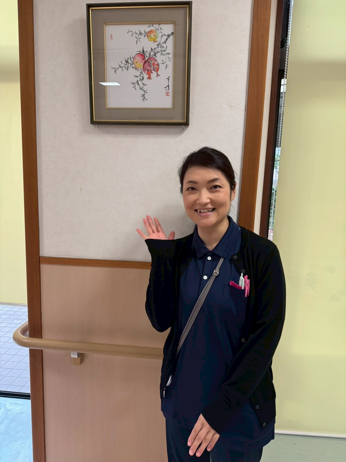社会福祉法人 若草会 太平の里デイサービスセンターの主任、岡本直美（おかもと なおみ）さんが笑顔で手を振る様子。濃紺の制服を着用し、胸ポケットにはペンが差されている。背景にはザクロの絵画と中国書道の額が飾られ、赤い飾りや観葉植物が彩りを添える。明るい室内で、親しみやすく温かな雰囲気が伝わる一枚。