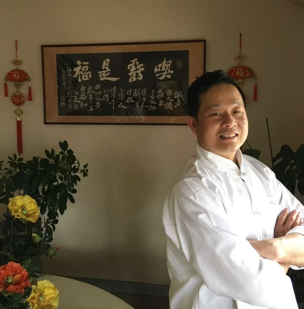 中国・天津出身の中華料理人、王 全信(ワン・チェンシン)さんのポートレート。白い伝統的な料理人の装いで腕を組み、笑顔を見せる姿が印象的。背景には中国書道の額や赤い飾りが配され、文化的な雰囲気と料理人としての誇りが感じられる一枚。