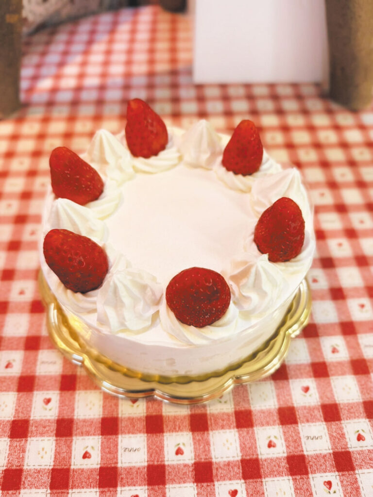 「フランス菓子マリークレール」のバースデーケーキ。金色の台座に乗せられた丸型ケーキには、白いホイップクリームの絞りと新鮮なイチゴが交互に飾られ、華やかで上品な仕上がり。背景の赤と白のチェック柄テーブルクロスにはハートやイチゴの模様があしらわれ、誕生日の特別感と可愛らしさを演出している。