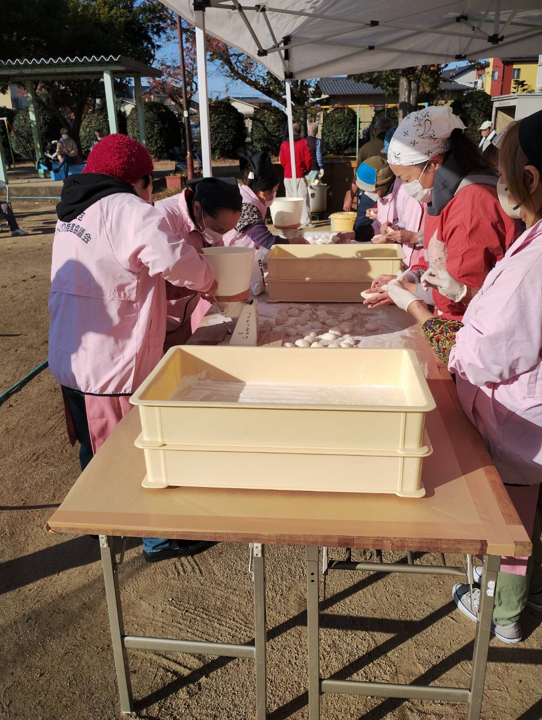 畑中餅つき大会の風景。白いテントの下で、参加者たちが黄色いトレーに並べられた白い丸餅を手際よく成形している。エプロンや手袋、帽子を着用し、衛生的に作業を進める様子が印象的。背景には「生活委員会」や「こども会」の表示があり、地域の世代を超えた交流と伝統行事の温かさが感じられる。