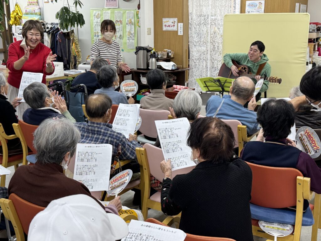 昭和の懐メロ音楽会の一幕。高齢者施設の室内で、ギターを奏でる男性を中心に、参加者が歌詞カードや「Happy!」と書かれたうちわを手に歌を楽しむ様子が写されている。笑顔で拍手を送るスタッフや、手振りで盛り上げる女性の姿もあり、温かく賑やかな雰囲気が漂う。壁には「クッキー会」などの掲示があり、地域交流と音楽を通じた心のふれあいが感じられる。