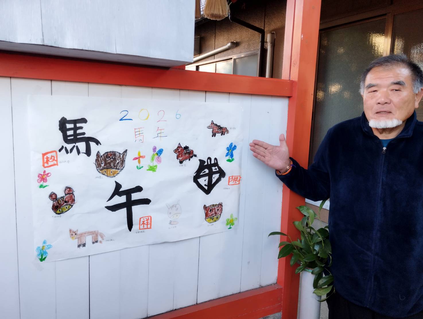 大分市荏隈大石町にある若宮八幡社で撮影された一枚。交通ボランティアとして活動する原さんが、壁に掲示された大きな書の作品の前に立っている。作品には「馬年」「2026」などの文字と、南大分の子どもたちが描いた動物や花のイラストが色鮮やかに並び、干支をテーマにした地域参加型の創作であることが伝わる。神社の静かな空間に、地域の思いと子どもたちの表現が調和した温かみのある場面。
