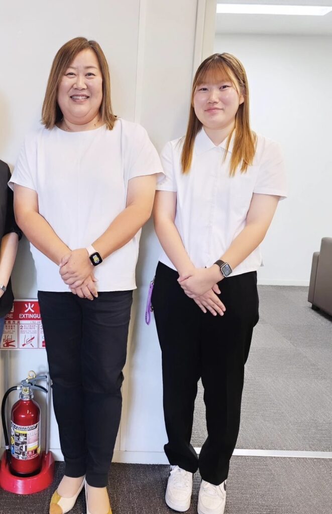 NPO法人子育て応援ワクワクピースの漆間文代さん（左）と、娘であり代表理事でもある漆間一夏さん（右）のツーショット。白いトップスと黒のパンツを揃えて着用し、落ち着いた室内で並んで立つ姿が写されている。背景には消火器や案内表示があり、活動拠点の一角で撮影されたと見られる。親子であり、地域の子育て支援を牽引する存在としての信頼感と温かさが伝わる一枚。