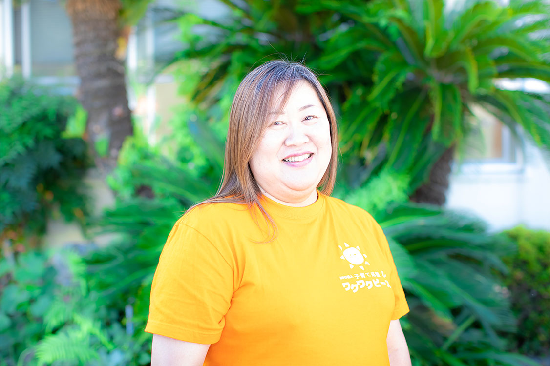 NPO法人子育て応援ワクワクピースの漆間文代さんの写真。緑豊かな屋外で、オレンジ色のTシャツを着て笑顔を見せている。Tシャツには ワクワクピースのロゴが書かれている。背景の植物と晴れやかな表情が、地域に根ざした温かい活動の雰囲気を伝えている。