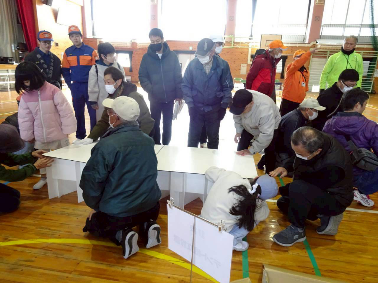 南海トラフ地震を想定した避難所開設・受入訓練の様子。体育館に地域住民や関係者が集まり、マスク姿で防寒着を着た参加者たちが、簡易ベッドを囲んで組み立て作業を行っている。職員やボランティアの姿も見られ、寒い時期の訓練らしい緊張感と協力体制が伝わる場面。