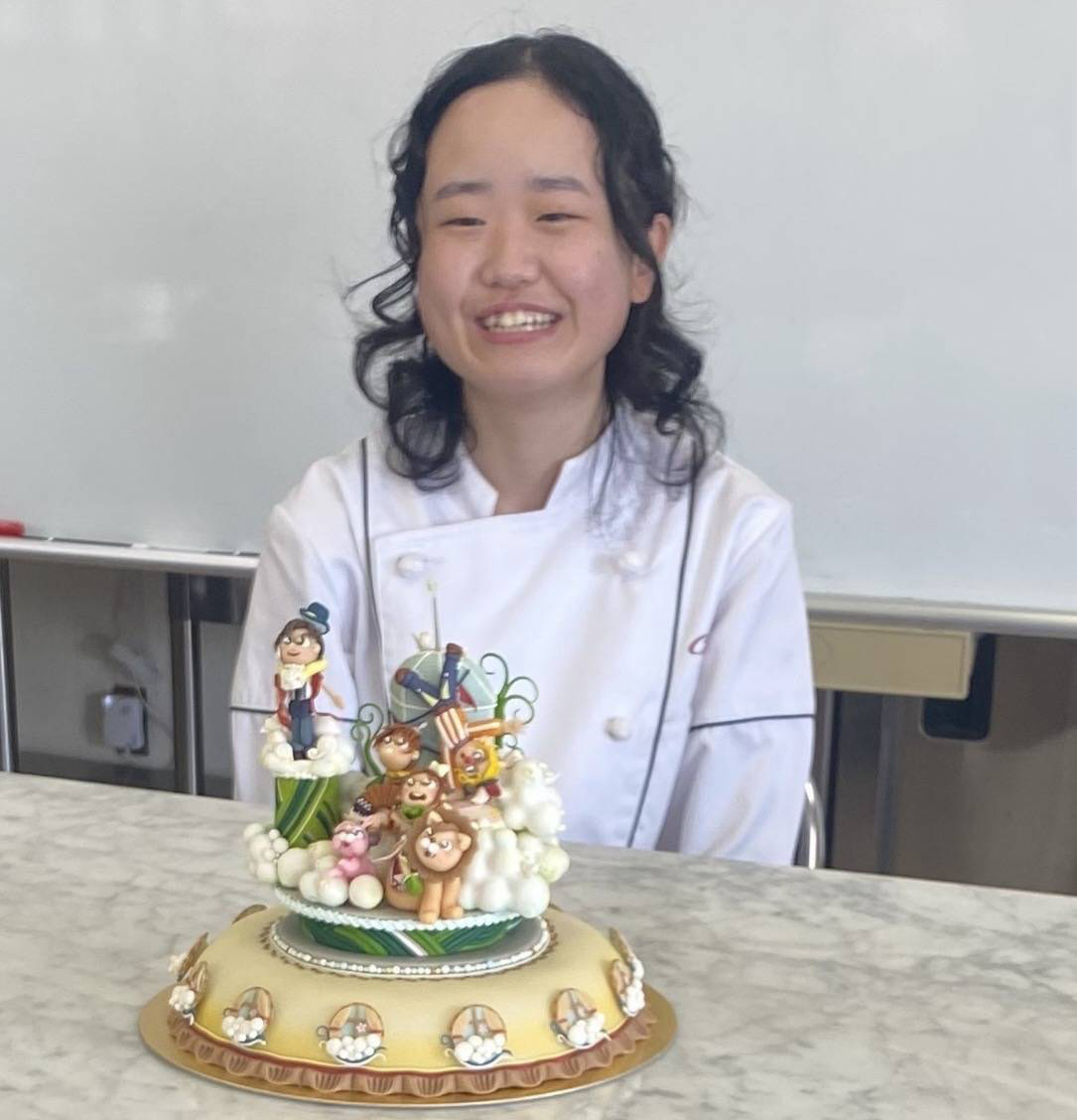 智泉福祉製菓専門学校 製菓製パン学科2年生の惣川日和（そうかわ ひより）さんが、2025年ジャパン・ケーキショーのマジパン・エコール部門で銀賞を受賞した際の写真。白衣姿で作品の後ろに座り、サーカスをテーマにしたと思われる色鮮やかなマジパン細工のケーキが手前に置かれている。ライオンなどの動物、テントや風船など細部まで作り込まれた装飾が並び、技術力と世界観の豊かさが伝わる構図。