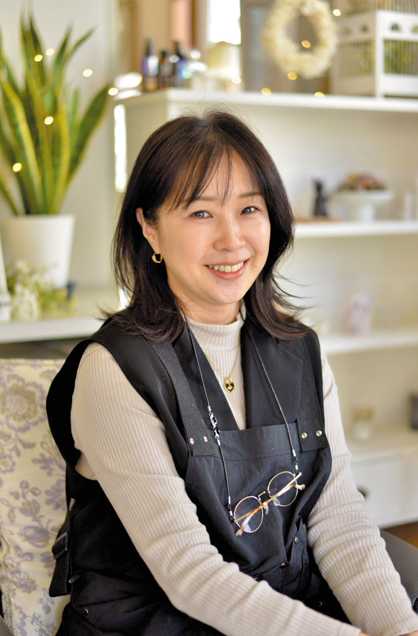 lily of the valley（リリーオブザバレー）代表の佐々木見和（ささき みわ）さんの写真。明るい室内で椅子に座り、柔らかな笑顔を向けている。淡い色のトップスに黒いエプロンを重ね、胸元には眼鏡チェーンとハート型のペンダントを身につけている。背後には観葉植物や棚に並ぶ雑貨があり、落ち着いた温かみのある空間が、サロンやワークスペースとしての雰囲気を伝えている。