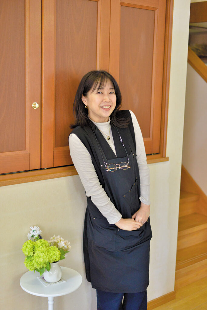 lily of the valley（リリーオブザバレー）代表の佐々木見和（ささき みわ）さんの写真。室内で木製キャビネットの前に立ち、柔らかな笑顔を向けている。淡い色のトップスに黒いエプロンを重ね、胸元には眼鏡チェーンが下がっている。左側には白い花瓶に生けられたグリーンと白の花が置かれ、背後には木の階段が見える。落ち着いた温かみのある空間が、サロンや活動拠点としての雰囲気を伝えている。