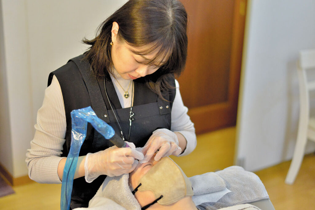 lily of the valley（リリーオブザバレー）代表の佐々木見和（ささき みわ）さんが、施術用ベッドに横たわる利用者に対して、専用のハンドツールを使って丁寧に施術を行っている様子。利用者は目元を覆うタオルを当ててリラックスした状態で、佐々木さんは手袋を着用し、衛生管理の整った環境で慎重に作業している。落ち着いた施術空間の中で、専門性と安心感が伝わる一場面。