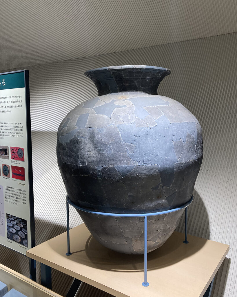 溝から出土した須恵器の大甕を復元した展示写真。多数の破片をつなぎ合わせた継ぎ目が表面に残り、丸みのある大きな甕が金属製の台座に載せられている。横には解説パネルが置かれ、考古資料としての価値と出土状況を伝える博物館展示の一場面。