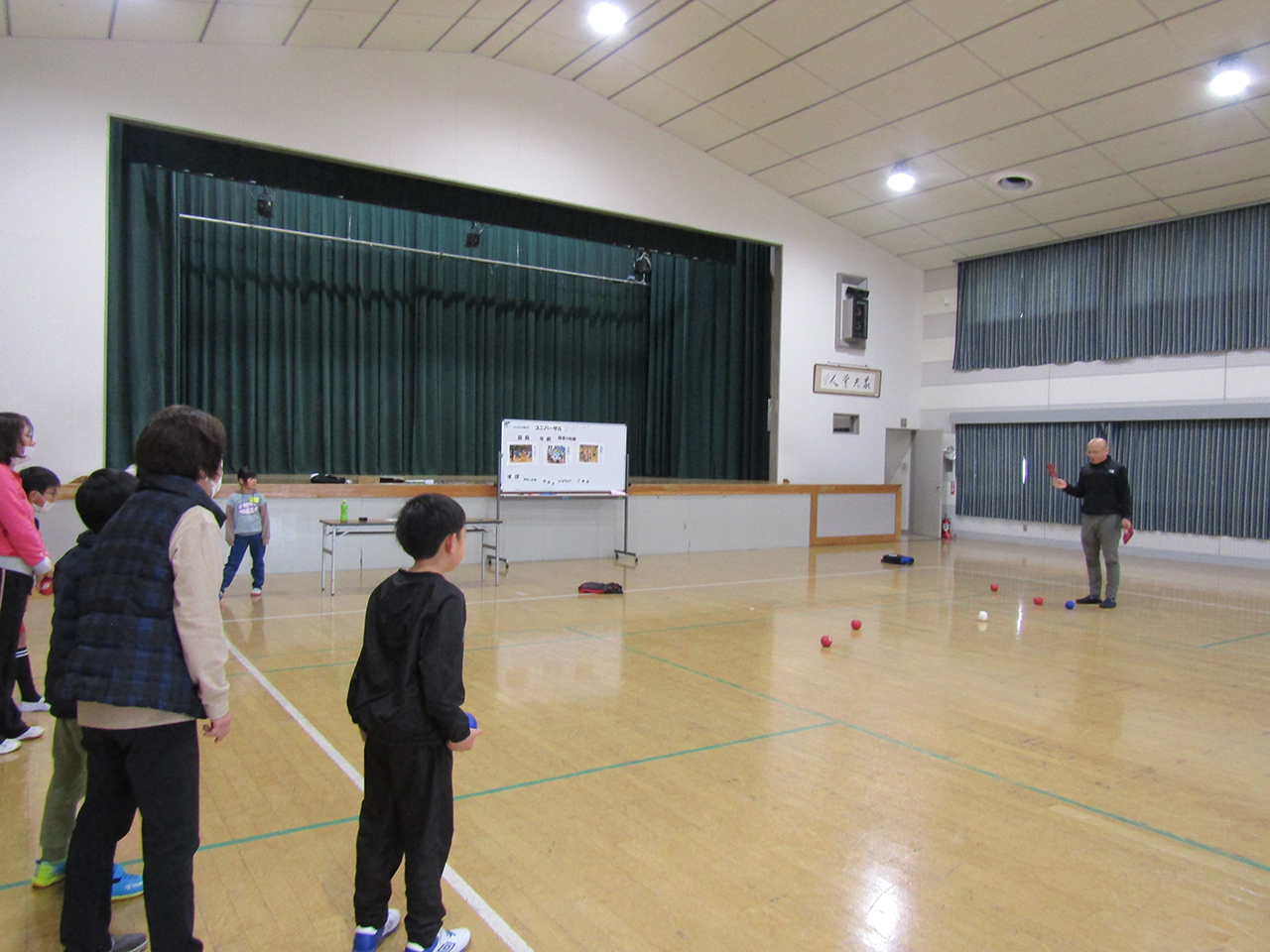 南大分公民館で行われた、4・5・6年生を対象としたボッチャ体験学習の様子。体育館の床に赤・青・白のボールが広がり、指導者が子どもたちに向けて投げ方を説明している。参加児童たちはコートの端に並んで見守り、ステージや掲示物が背景に見える、地域学習らしい和やかな雰囲気の場面。