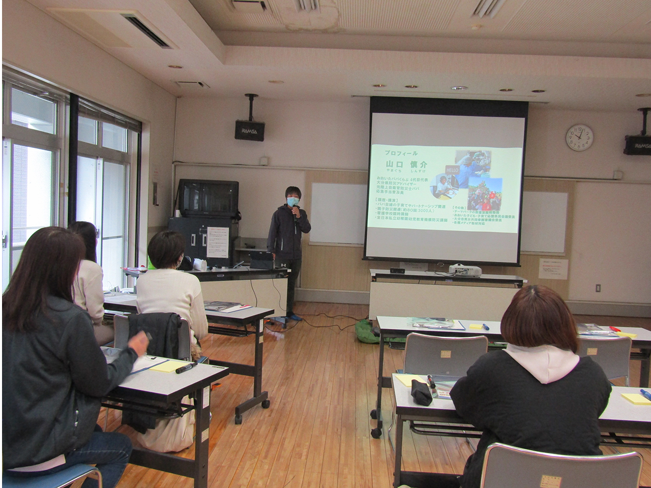 防災教室の様子。講師がマイクを持って前方に立ち、スクリーンに映したプロフィール資料を示しながら受講者に向けて説明している。教室内では参加者が机に着席して講義を聞いており、大きな窓から光が差し込む明るい学習環境が広がっている。地域の防災意識向上を目的とした講座の雰囲気が伝わる場面。