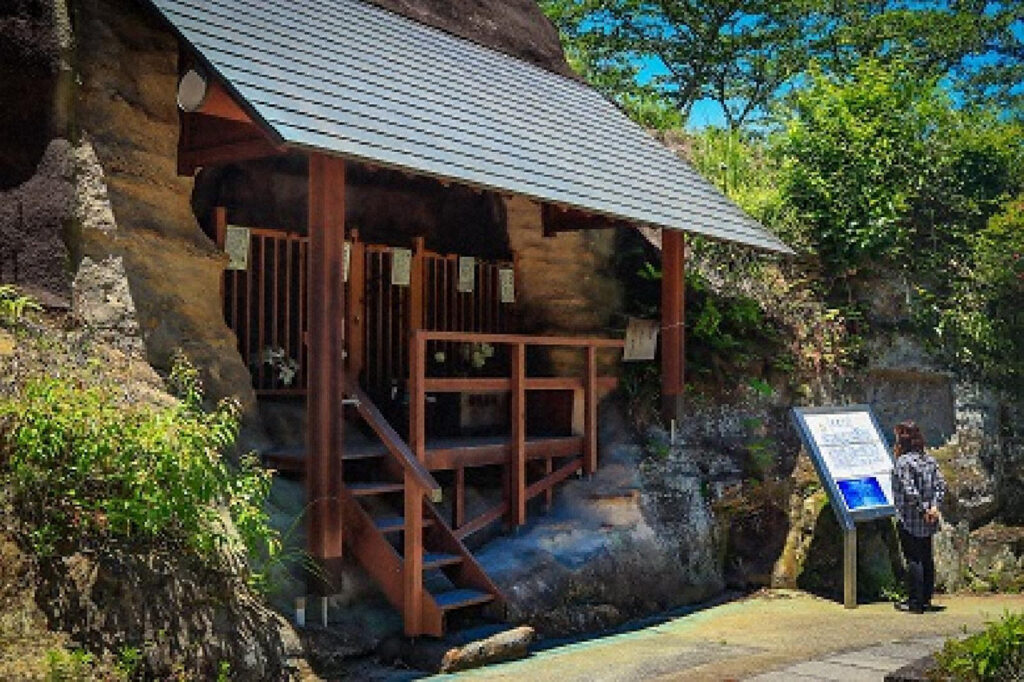 岩肌に造られた高瀬石仏を覆うように建てられた小さな覆屋の前景。木製の柱と金属屋根の下には石仏を祀る空間があり、手前には参拝者向けの案内板を読む人物が立っている。背後には岩壁と緑が広がり、青空の下で自然と文化財が調和した現在の高瀬石仏の姿が写されている。