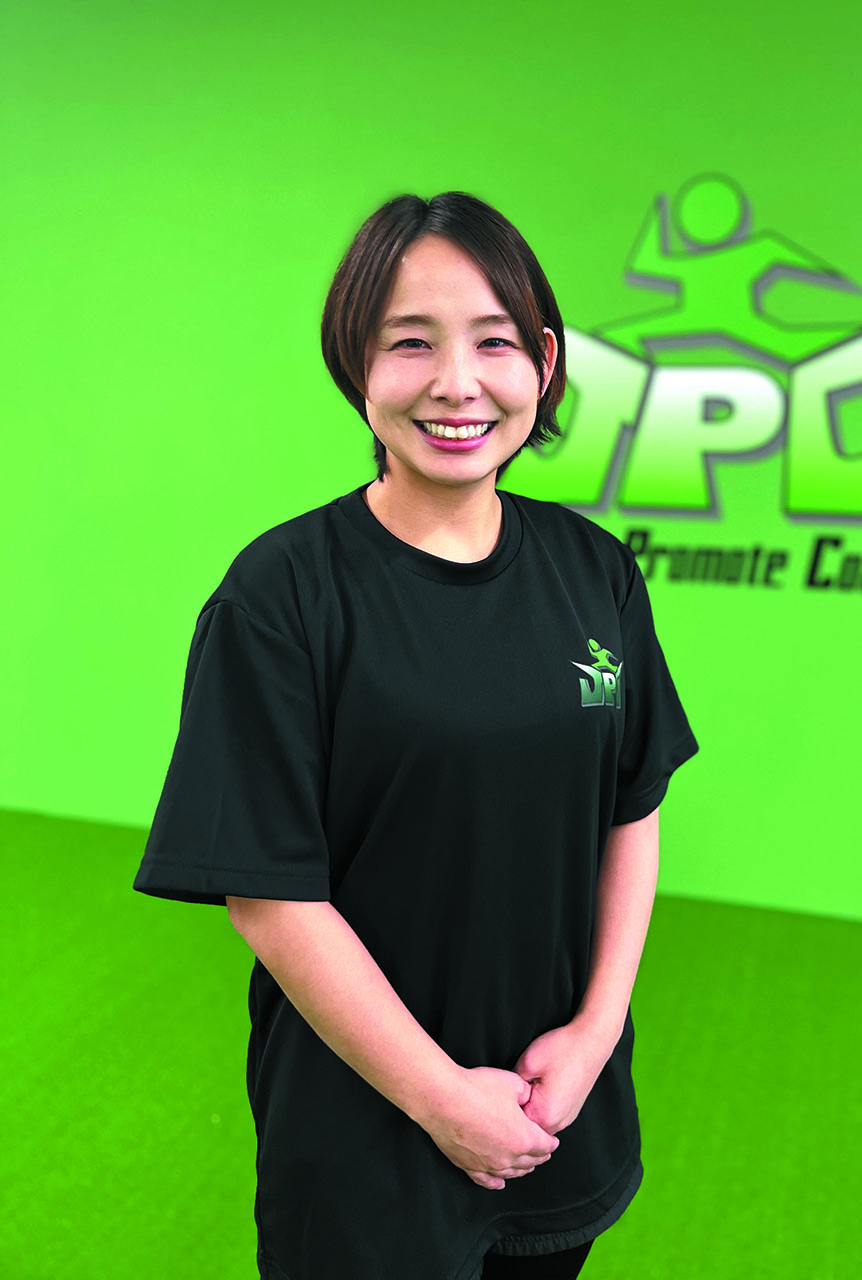 JPCスポーツ教室　大分古国府店「コーチ」の工藤 暢子さん。緑の壁にロゴの入った背景をバックに黒いトレーニングウェアを着て笑顔でこちらを見ています。