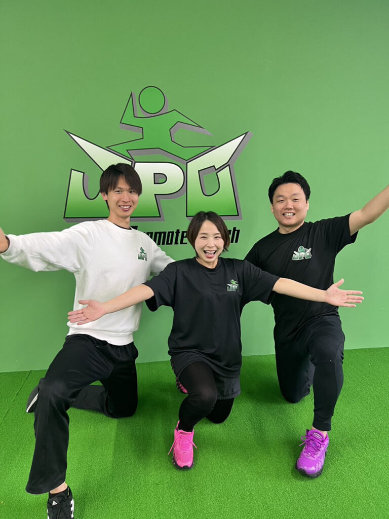 JPCスポーツ教室のスタッフ3人で楽しそうにポーズを決めています。緑の壁にロゴの入った壁をバックにポーズを決めています。