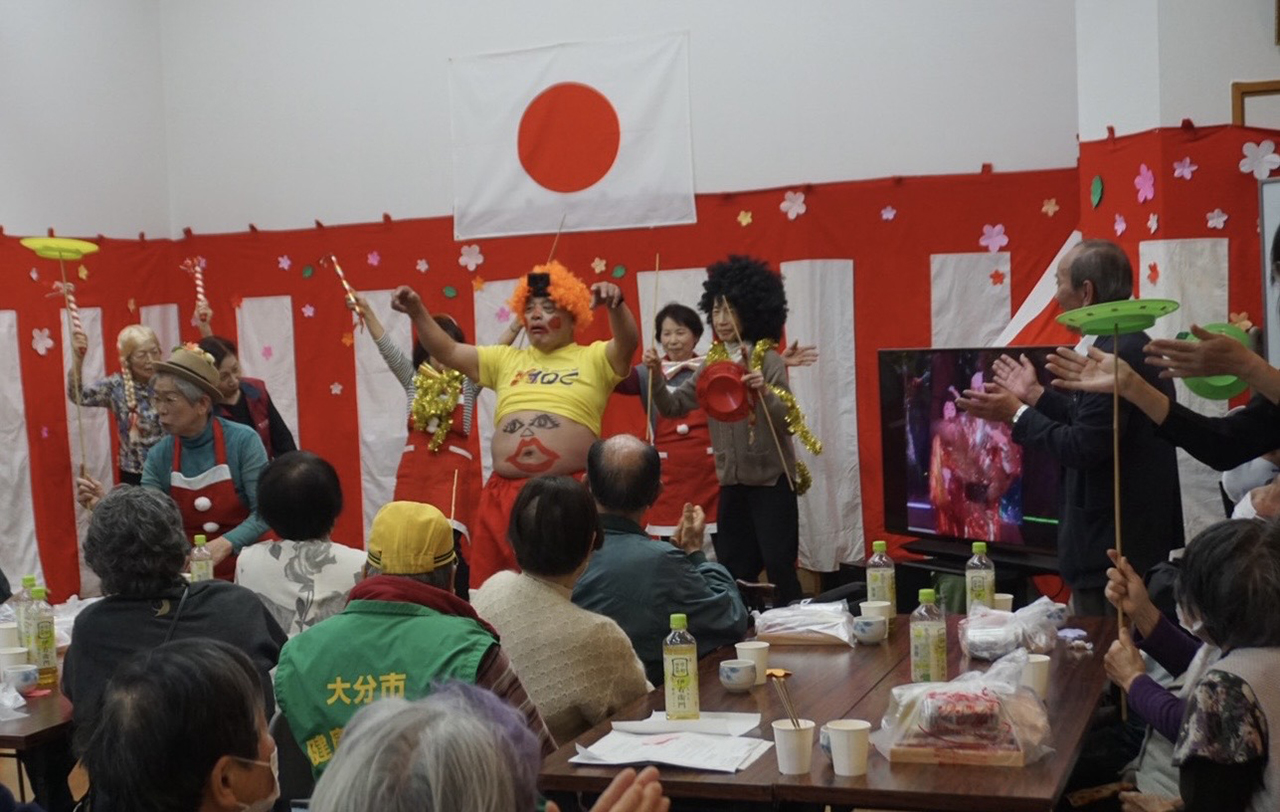 豊府社協・豊府民児協共催のお花見会の様子を写した写真。舞台前では、カラフルな衣装や小道具を身につけた出演者たちがパフォーマンスを披露し、客席では参加者が拍手をしながら楽しんでいる。背景には紅白の飾りや大きな日の丸が掲げられ、テーブルには飲み物や資料が置かれた、地域の交流イベントらしいにぎやかな雰囲気が伝わる。